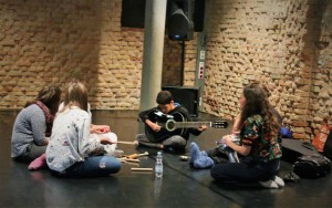 Kinder sitzen auf den Boden und schauen zu einem Jungen, der sitzend Gitarre spielt