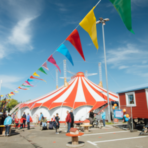 Eine bunte Wimpelkette hängt unter freiem Himmel bei Sonnenschein auf dem Circus Fantasia Platz. Im Hintergrund stehen Menschen und das große Circuszelt.