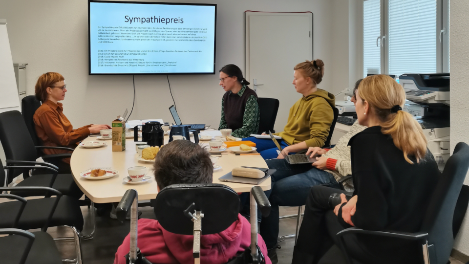 Die Jury des DIALOGE sitzen um eine Kaffeetafel herum, in Hintergrund ist mit einem Beamer eine Powerpoint an eine Leinwand projiziert