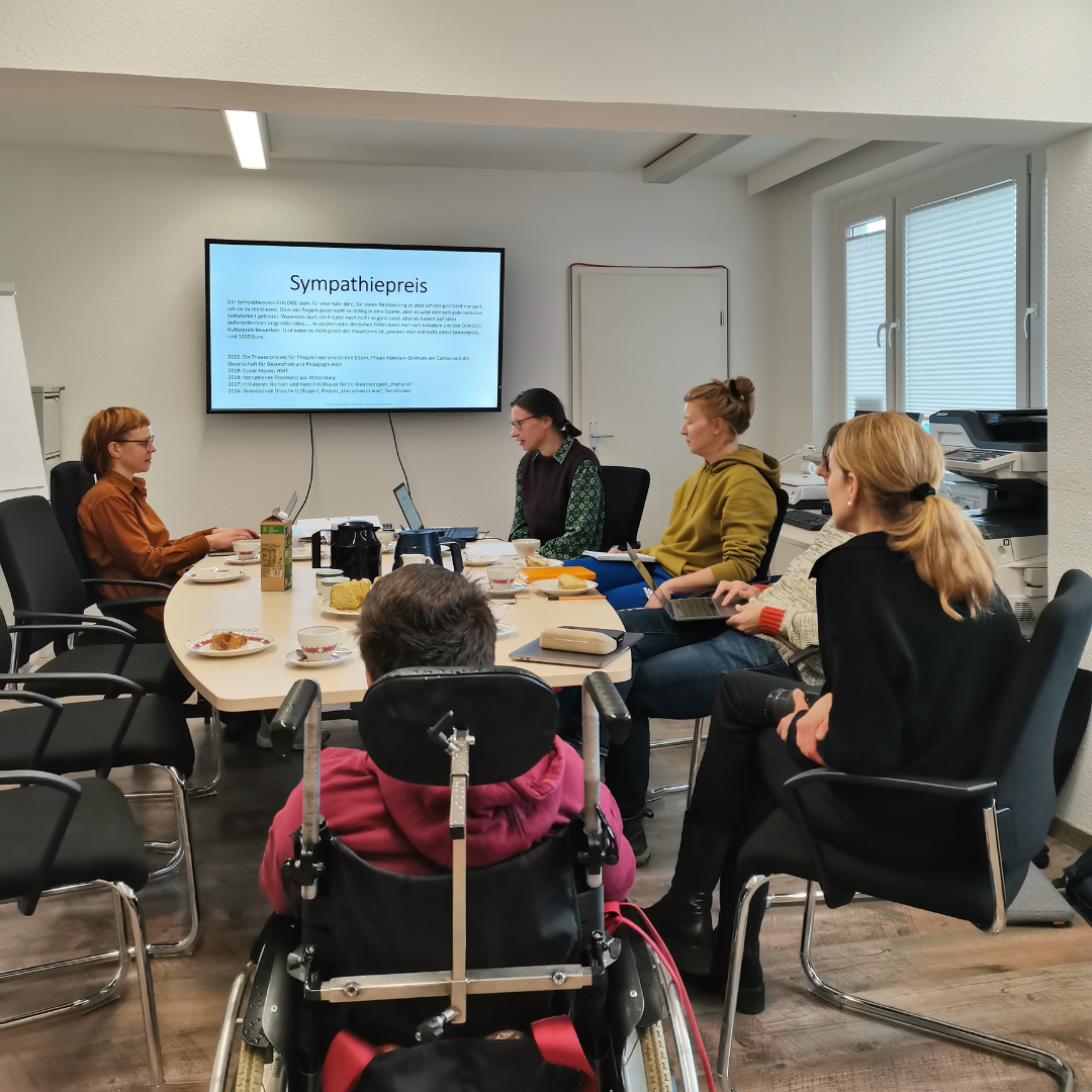 Die Jury des DIALOGE sitzen um eine Kaffeetafel herum, in Hintergrund ist mit einem Beamer eine Powerpoint an eine Leinwand projiziert