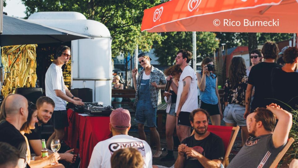 Zu sehen ist ein DJ und verschiedene Menschen, die tanzen und auf Stühlen unter freiem Himmel sitzen