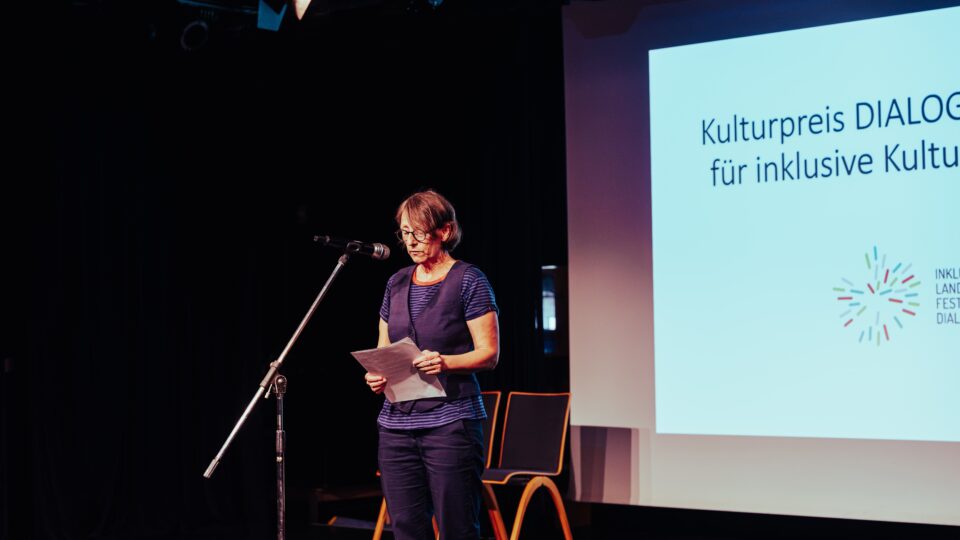 Eine Frau steht vor einer Leinwand mit der Aufschrift "Kulturpreis DIALOGE" und verkündet den/die Gewinner*in des DIALOGE-Hauptpreises